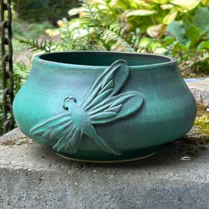 May include: A round, teal-colored ceramic bowl featuring a raised, sculpted dragonfly design on its exterior. The bowl has a smooth, matte finish with subtle variations in color, showcasing a wide opening and a curved, tapered base profile.