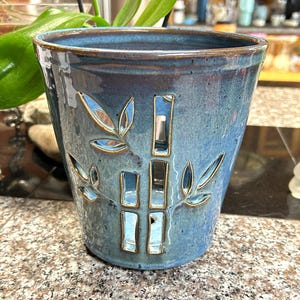 May include: A tapered ceramic planter with a glossy blue glaze and a speckled finish. The exterior features cut-out designs of bamboo stalks and leaves, with the edges of the cut-outs highlighted in a contrasting brown tone.