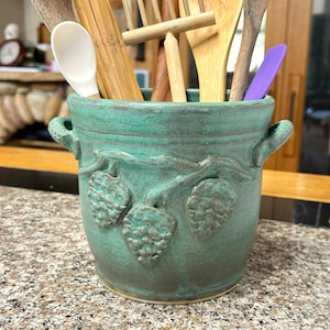 May include: A teal ceramic utensil holder with a textured surface and embossed grape vine design. The holder has two handles and is filled with wooden spoons, a white spoon, and a purple spatula. A decorative kitchen item.