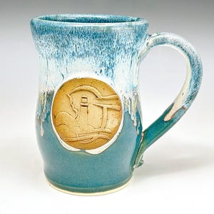 May include: A handcrafted ceramic mug with a coastal theme. The mug features a teal base, a light blue top with a dripped glaze effect, and a large handle. A circular emblem with a lighthouse design is centered on the mug.