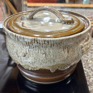 May include: A handmade ceramic casserole dish with a lid. The dish is a mix of brown, beige, and white with a textured, dripped glaze. It has a handle on the lid and two side handles. The dish is approximately 7 inches in diameter.