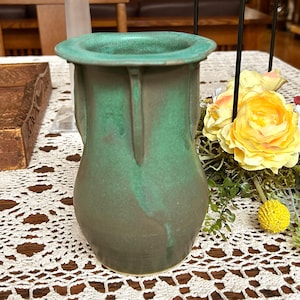 May include: A handcrafted ceramic vase with a green and brown glaze. The vase has a wide, flared rim and two vertical handles. The vase is approximately 8 inches tall and is sitting on a white lace tablecloth.
