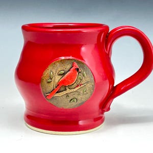 May include: A bright red ceramic mug featuring a circular medallion on the front. The medallion displays a raised, detailed relief of a red cardinal bird perched on a branch with small leaves against a textured, earthy brown background.