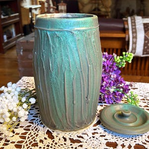 May include: A handcrafted, cylindrical ceramic vase with a lid. The vase has a textured, vertical pattern in shades of teal and gray-green. The lid is round with a small knob. The vase is displayed on a white lace tablecloth, with flowers in the background.