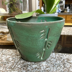 May include: A handcrafted, green ceramic planter with a textured surface. The planter features cut-out bamboo leaf designs. The interior is a darker green, and the rim has a darker, weathered appearance. The planter is suitable for indoor plants.