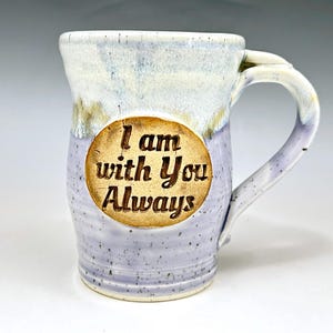May include: A handmade ceramic mug with a light blue and white glaze. The mug features a brown, circular emblem with the engraved text "I am with You Always". The handle is a curved, off-white color.