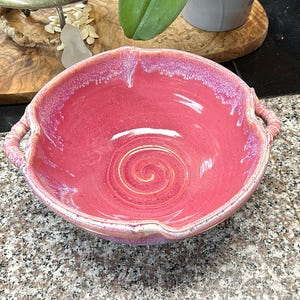 May include: A handmade, ceramic bowl in a vibrant pink hue, featuring a unique spiral design at the base. The bowl has two handles and a textured rim. This decorative piece is ideal for serving or display.