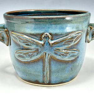 May include: A ceramic bowl with two side handles, featuring a glossy blue and brown glaze. The exterior is decorated with a prominent, embossed dragonfly motif that spans the front, showcasing detailed wing patterns and a slender body.