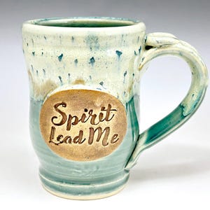 May include: A ceramic mug with a glossy glaze in shades of teal, blue, and cream. The mug features a prominent circular medallion on the front with the embossed text Spirit Lead Me. It has a sturdy handle and a slightly flared rim with drip details.