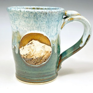 May include: A handmade ceramic mug with a mountain design. The mug features a light blue and green glaze with a brown mountain scene in a circular inset. The handle is a curved design with a matching glaze.