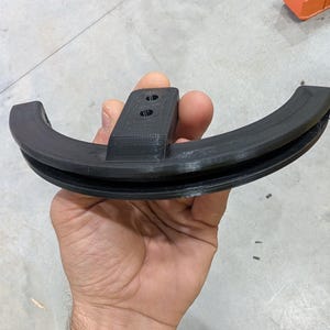 May include: A black, 3D-printed, semi-circular object with a central rectangular section featuring two holes. The object has a grooved design along its curved edges. The item is held in a hand.
