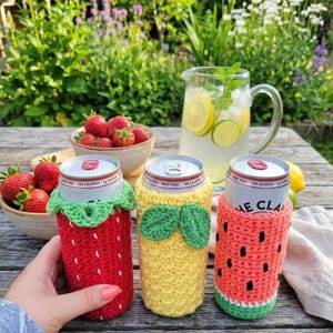 May include: Three canned beverages in handmade crochet cozies shaped like a strawberry, pineapple, and watermelon. The red strawberry cozy has a green leaf trim. The yellow pineapple cozy has green leaves. The watermelon cozy is red with black seeds and a green rind.