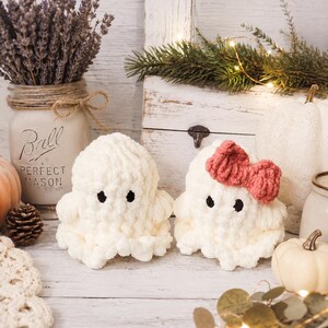 May include: Two small, plush, cream-colored crocheted ghost figures with black embroidered eyes. One ghost features a textured, ruffled base and a rounded top, while the other identical ghost is adorned with a dusty rose-colored crocheted bow.