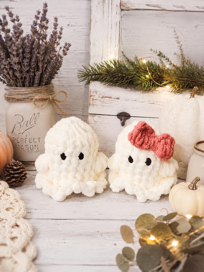 May include: Two small, plush, cream-colored crocheted ghost figures with black embroidered eyes. One ghost features a textured, ruffled base and a rounded top, while the other identical ghost is adorned with a dusty rose-colored crocheted bow.