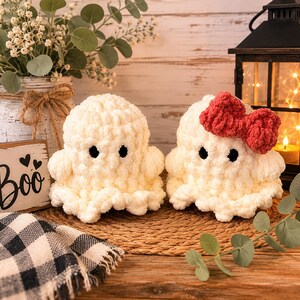 May include: Two handmade crochet ghosts made of soft white yarn with black safety eyes. One ghost features a prominent red yarn bow attached to its head. The ghosts have a rounded, plush texture and sit side by side on a woven circular mat.