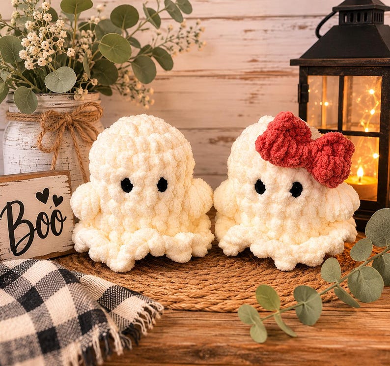 May include: Two handmade crochet ghosts made of soft white yarn with black safety eyes. One ghost features a prominent red yarn bow attached to its head. The ghosts have a rounded, plush texture and sit side by side on a woven circular mat.