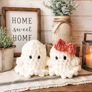 May include: Two white crocheted ghost plushies with black eyes, one featuring a small red crocheted bow on its head. The plushies are made of a soft, chunky yarn and have a rounded, textured appearance with ruffled edges at the base.