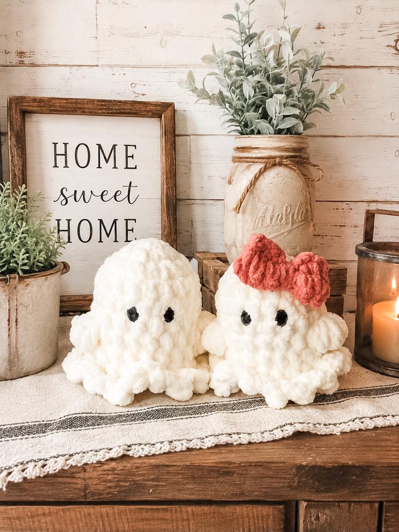 May include: Two white crocheted ghost plushies with black eyes, one featuring a small red crocheted bow on its head. The plushies are made of a soft, chunky yarn and have a rounded, textured appearance with ruffled edges at the base.