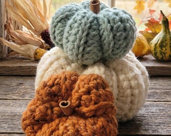 May include: A stack of three crocheted pumpkins in sage green, cream, and burnt orange. Each pumpkin features a thick, ribbed yarn texture and a small, brown cinnamon stick stem protruding from the center top of the rounded, soft form.