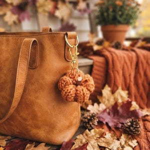 May include: A tan leather tote bag with a gold-toned metal lobster clasp keychain attached to the side ring. The keychain features a small, round, orange crocheted pumpkin with a short, light-colored wooden stem attached to the top.