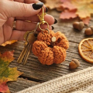 May include: A crocheted orange pumpkin keychain featuring a small wooden stem, attached to a gold-toned metal lobster clasp and a split ring holding two silver-colored metal keys. The pumpkin is crafted with textured yarn in a rounded, ribbed shape.