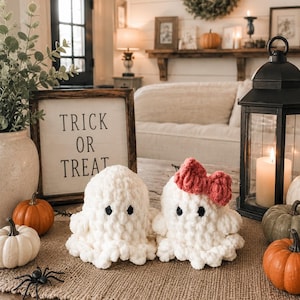 May include: Two white crocheted ghost plushies with black eyes, one featuring a small red bow on its head. They are positioned next to a wooden sign with the text TRICK OR TREAT, small orange and white pumpkins, and a black plastic spider.