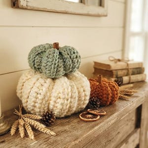 May include: Three crocheted pumpkins in sage green, cream, and burnt orange, each topped with a small brown stem. The pumpkins are arranged with dried wheat stalks, pinecones, and dried orange slices on a rustic wooden mantelpiece.