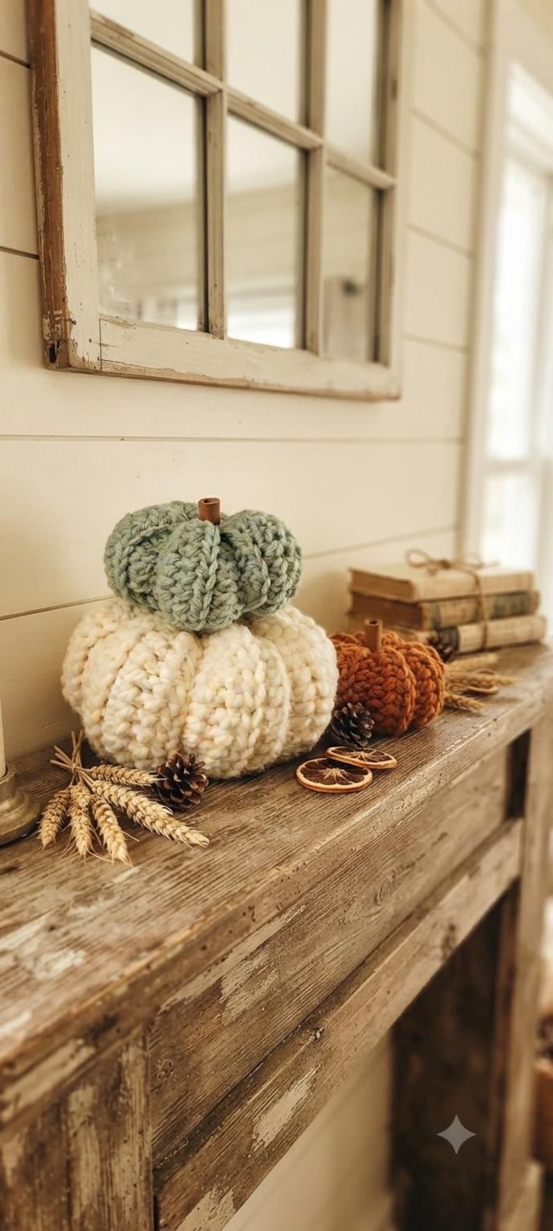 May include: Three crocheted pumpkins in sage green, cream, and burnt orange, each topped with a small brown stem. The pumpkins are arranged with dried wheat stalks, pinecones, and dried orange slices on a rustic wooden mantelpiece.