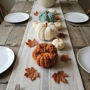 May include: A dining table setting featuring a light gray linen runner decorated with three chunky knit pumpkins in sage green, cream, and burnt orange, accompanied by small white gourds and scattered dried brown maple leaves.