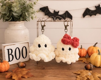 May include: Two white crochet ghost keychains with black plastic eyes, one featuring a red crochet bow, attached to bronze-colored lobster clasps. The ghosts are displayed on a metal stand, with a wooden sign reading BOO nearby.