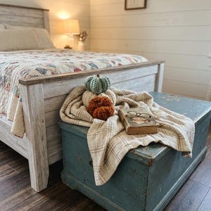 May include: A stack of three crocheted pumpkins in sage green, cream, and burnt orange resting on a beige plaid throw blanket atop a blue wooden chest, accompanied by a vintage hardcover book and a pair of wire-rimmed reading glasses.