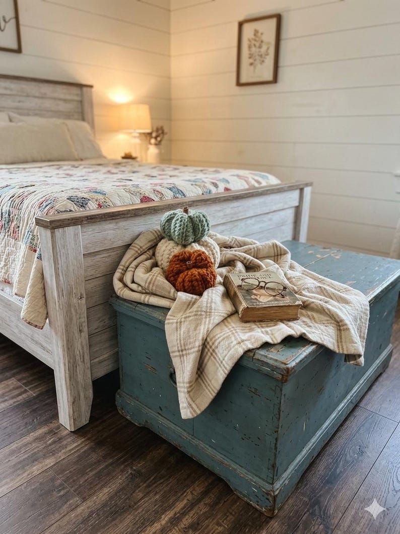May include: A stack of three crocheted pumpkins in sage green, cream, and burnt orange resting on a beige plaid throw blanket atop a blue wooden chest, accompanied by a vintage hardcover book and a pair of wire-rimmed reading glasses.