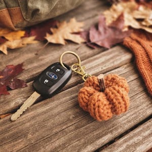 May include: A keychain featuring a small, crocheted orange pumpkin with a brown stem attached to a brass-toned metal lobster clasp and ring, connected to a black plastic Subaru car key fob with three buttons and a silver metal key blade.
