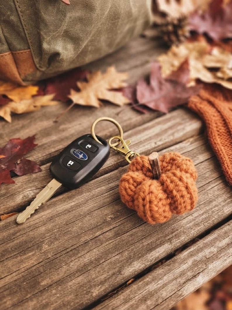 May include: A keychain featuring a small, crocheted orange pumpkin with a brown stem attached to a brass-toned metal lobster clasp and ring, connected to a black plastic Subaru car key fob with three buttons and a silver metal key blade.