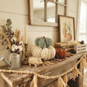 May include: A rustic mantel display featuring three chunky knit pumpkins in sage green, cream, and burnt orange. The arrangement includes a galvanized metal watering can with dried cotton and wheat, a brass candle holder, and a wooden bead garland.