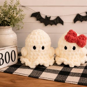 May include: Two handmade crochet plush ghosts made from soft white yarn. Both ghosts feature small black circular eyes. One ghost is plain, while the other is adorned with a bright red crocheted bow attached to the side of its head. The texture is chunky.