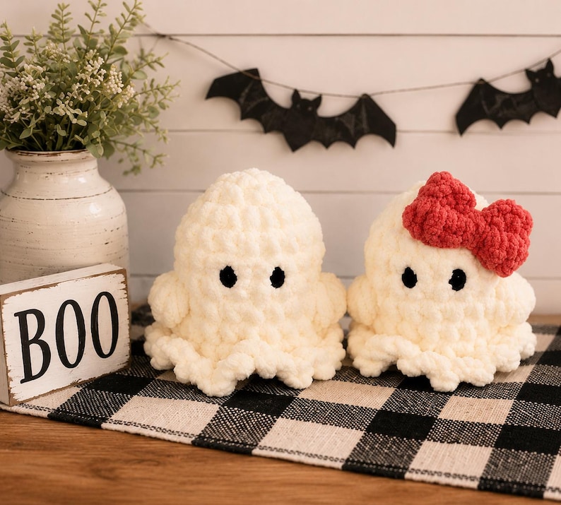 May include: Two handmade crochet plush ghosts made from soft white yarn. Both ghosts feature small black circular eyes. One ghost is plain, while the other is adorned with a bright red crocheted bow attached to the side of its head. The texture is chunky.