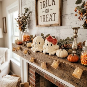 May include: Two white crocheted ghost plushies, one with a red bow, sit on a wooden mantel with a Trick or Treat sign, small pumpkins, jars of candy corn, a silver candlestick, and a string of burlap-covered fairy lights for Halloween decor.