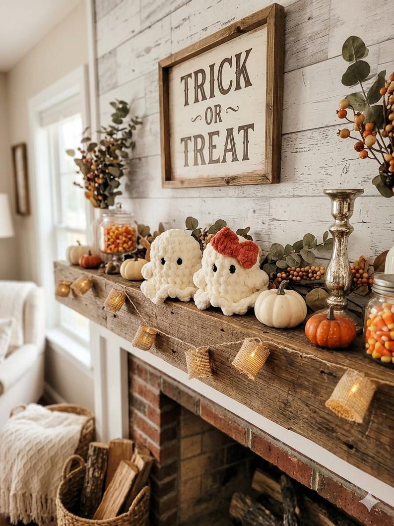 May include: Two white crocheted ghost plushies, one with a red bow, sit on a wooden mantel with a Trick or Treat sign, small pumpkins, jars of candy corn, a silver candlestick, and a string of burlap-covered fairy lights for Halloween decor.