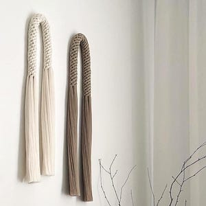 May include: A minimalist interior scene featuring a wooden table with decorative items. A textured white vase holds branches, a small white vase, and a black abstract sculpture. Two hanging wall decorations in beige and brown add to the neutral color palette.