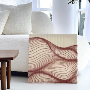 May include: A square canvas art piece featuring a cream-colored background with a repeating pattern of wavy, reddish-brown lines. The artwork is displayed in a bright, modern living room setting, next to a white sofa and a small wooden side table.