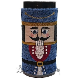 May include: A blue and red fabric cup cozy with an embroidered nutcracker design. The nutcracker has a white beard, blue eyes, and a black mustache.