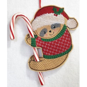 May include: A brown sloth wearing a red and green sweater and a Santa hat holds a candy cane. The sloth is a Christmas ornament.