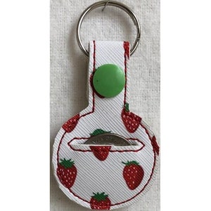 Op de afbeelding: Een witte stoffen sleutelhanger met een rood aardbeienpatroon. Hij heeft een groene knop en een klein vakje voor een munt. De sleutelhanger is bevestigd aan een zilveren sleutelring.