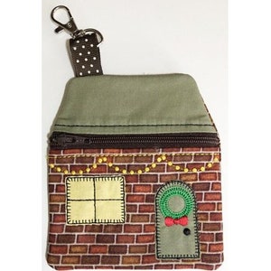 May include: A small, brown brick house-shaped zippered pouch with a green roof, a window, and a door with a wreath. The pouch has a brown and white polka dot strap with a silver clip.
