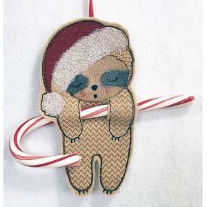 May include: A brown sloth wearing a red and white Santa hat holds a candy cane in its arms. The sloth is embroidered on a white fabric.