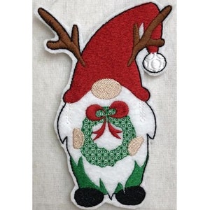 May include: Embroidered Christmas gnome with a red hat, brown antlers, and a green wreath with a red bow.