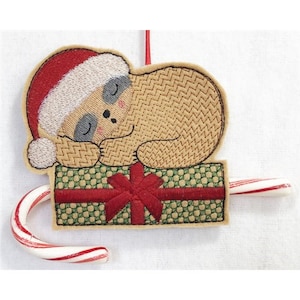 May include: Embroidered Christmas ornament featuring a sleeping sloth wearing a Santa hat. The sloth is resting on a green and red gift box, with a candy cane extending from the box. The ornament has a red hanging string.