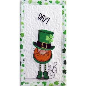 May include: Embroidered St. Patrick's Day wall hanging featuring a leprechaun with an orange beard, green hat, and boots. The white background has a quilted pattern and the word "DAY!" is written above the leprechaun. The border is green with shamrocks.