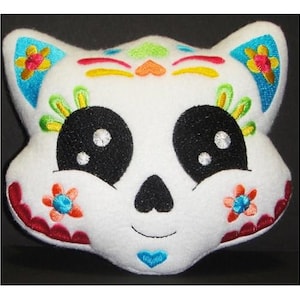 May include: White plush cat head with embroidered details in a Day of the Dead style. The cat has black eyes, a black nose, and a red mouth. The embroidery includes flowers, hearts, and swirls in various colors.